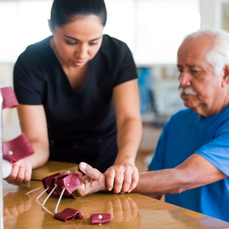 A senior male patient sitting and exercising his hand with a physical therapy device next to a occupational therapist.