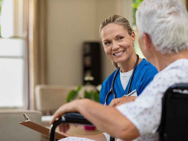 Friendly doctor examining health of patient sitting in wheelchair. Happy smiling nurse consulting disabled patient about treatment. Nurse caring about elder handicap man at home.